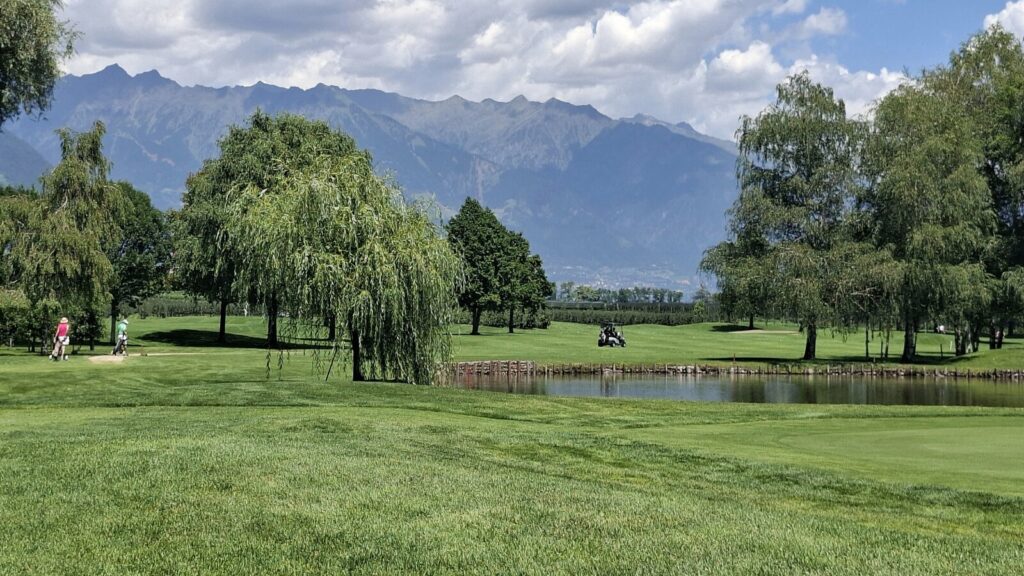 Teich auf dem Golfplatz Lana mit Golfspielern, Bäumen und Bergpanorama 